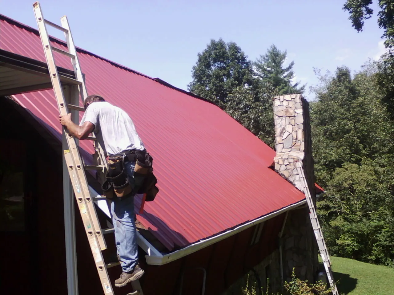 Roofer on a ladder beside a red metal roof and stone chimney — Soffit & Fascia Repair by Roofing Brothers in Westover